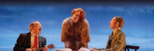 Three people at a table, two men sat on either side while a woman in a fur stole is stood up speaking